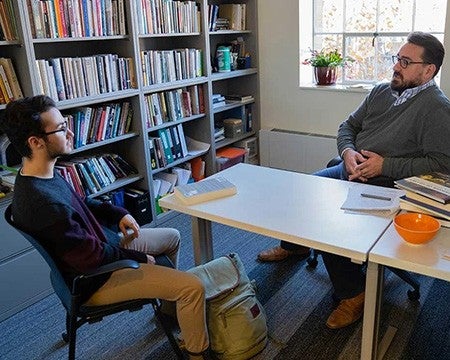 Male professor meeting with a bearded student in book-lined faculty office.