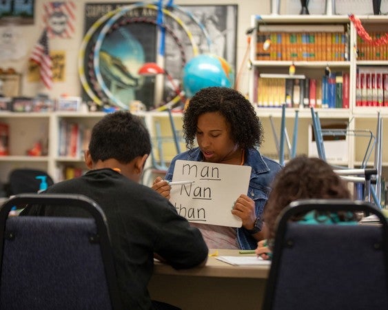 Holding a flash card, a woman helps two children with a language lesson in an elementary classroom.