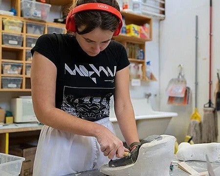 A student sculpting an object in clay on tabletop while wearing headphones.