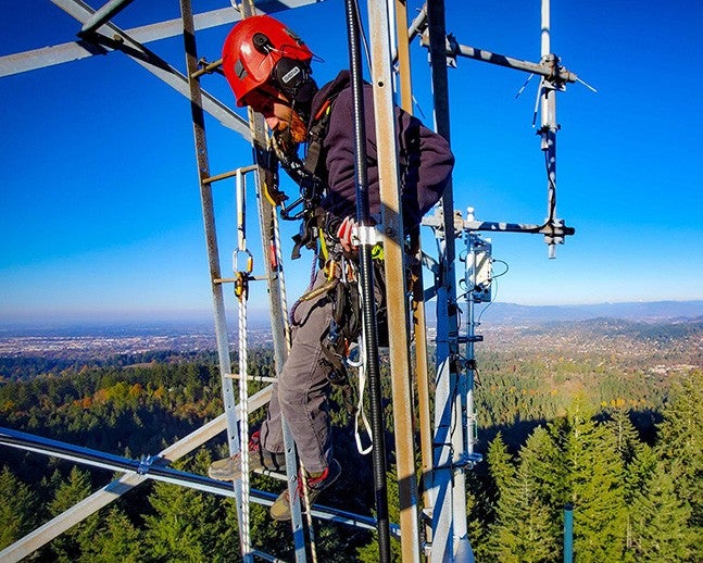 Student in a hardhat stands high above treetops in wildfire observation tower