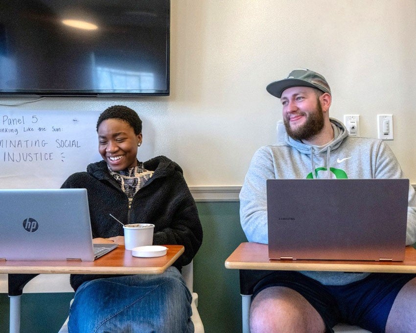 Two students smile and chat while sitting with their laptops on tables in front of them