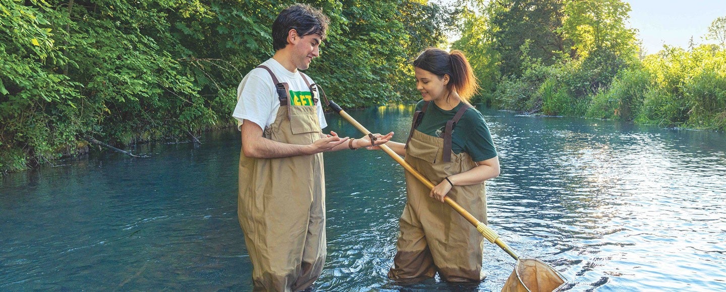 Two students standing in a river while wearing chest waders and holding a net during a course-related activity.